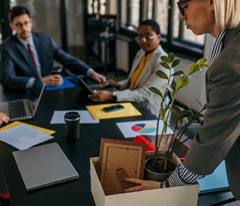Employee packing personal items into a box while colleagues discuss in the background, representing voluntary redundancy in the workplace.